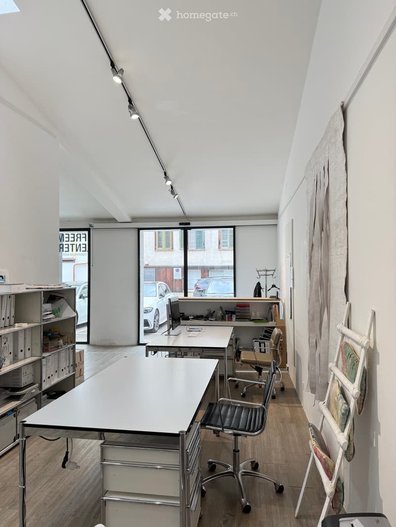 Modern office space with white walls, wooden flooring, and large windows. Features include a large white table, office chairs, a computer screen, shelves with office supplies, and a hanging lamp.