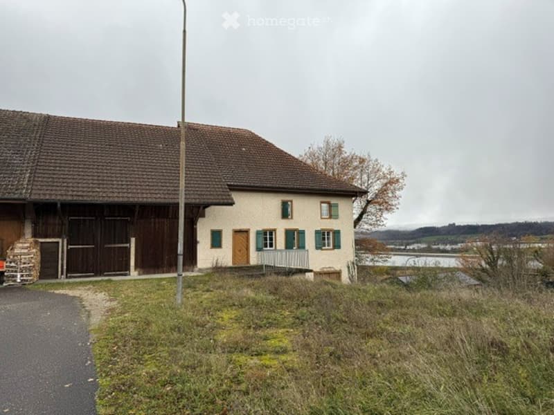 Traditional two-story house with a brown tiled roof, white walls, green shutters, and a wooden door. A wooden outbuilding is attached to the left side. The house is situated on a grassy slope overlooking a body of water.