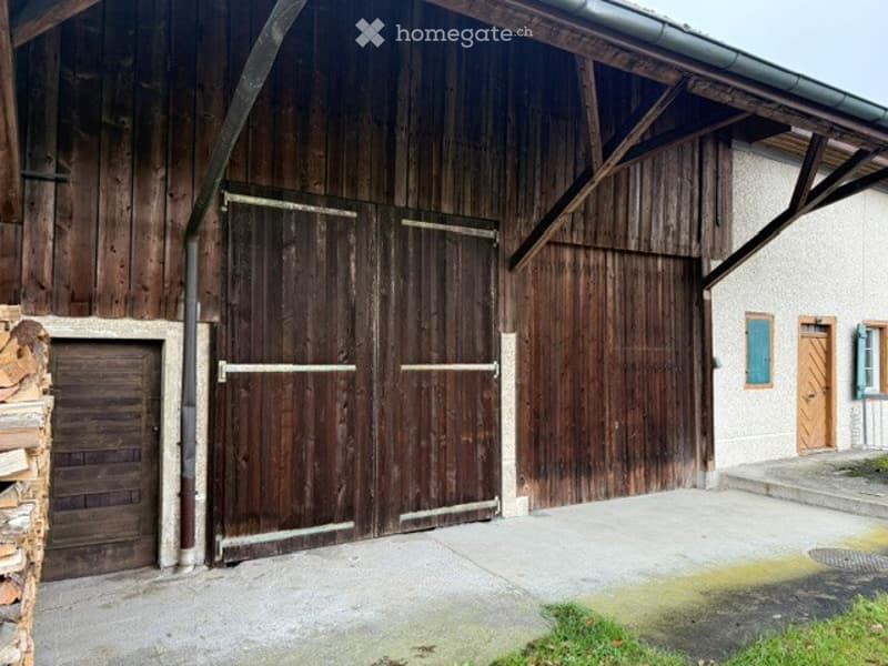Large wooden barn with two large garage doors and a small entrance door, adjacent to a house with blue shutters.