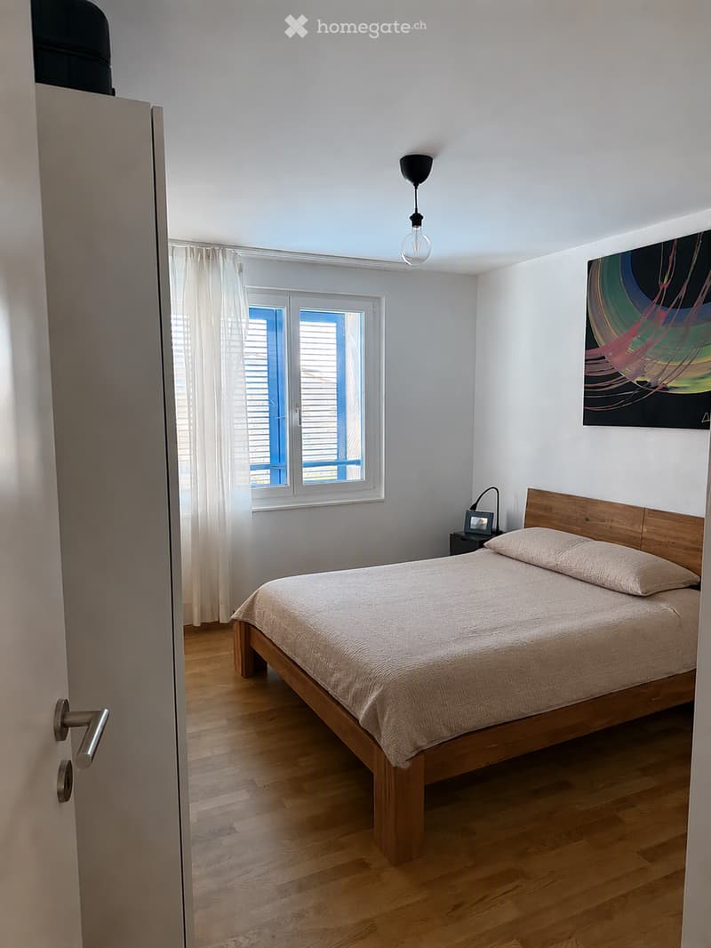 A modern bedroom with a wooden bed, nightstand, and a hanging pendant light. The room features light-colored walls, wooden flooring, and a large window with white curtains.