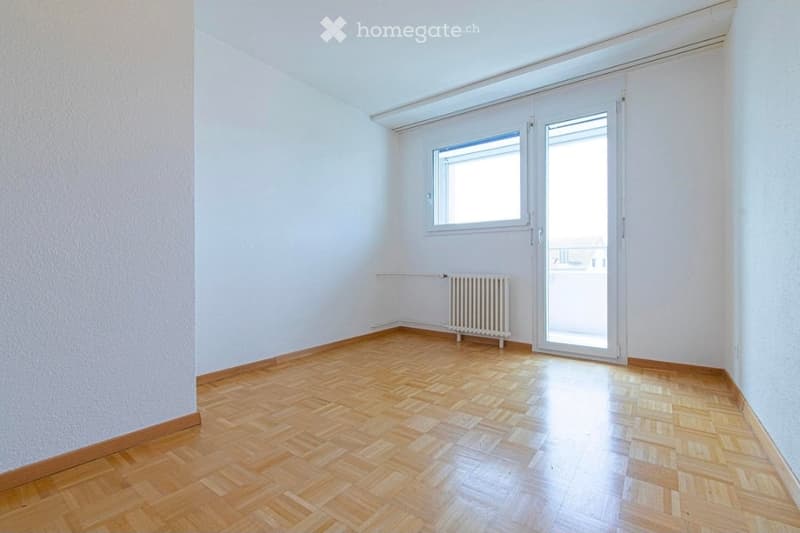 Empty room with wooden parquet flooring, white walls, a radiator, a window, and a glass door leading to a balcony.