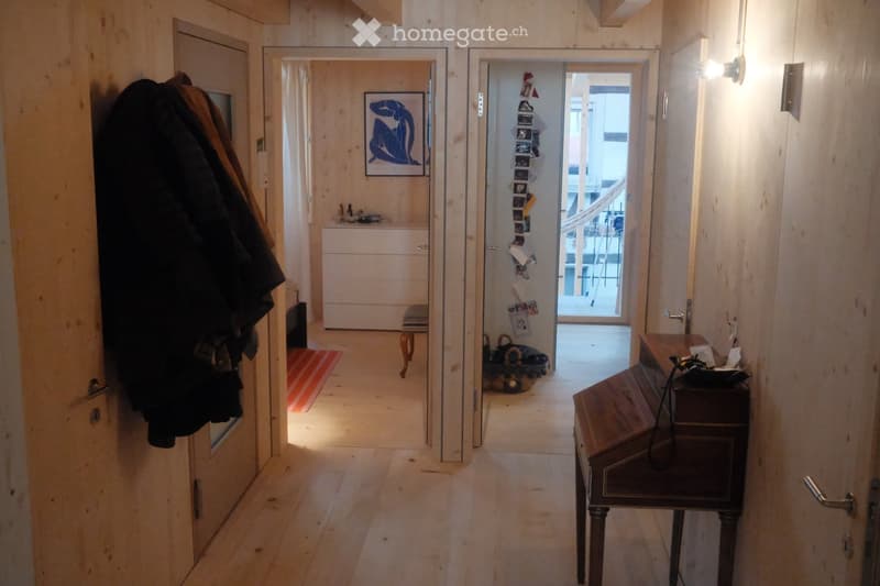 A narrow wooden hallway with multiple doors, a small bench, a coat rack, and a small table with items on it. Natural light streams in from the end of the hallway.