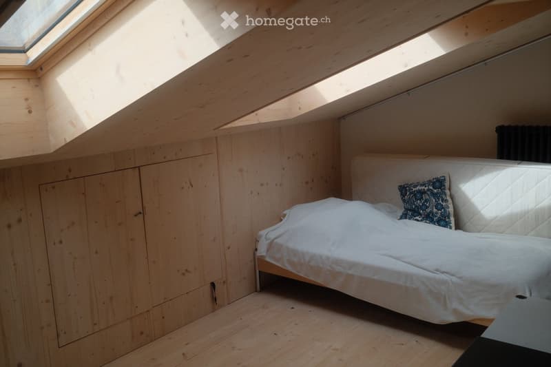 Cozy attic bedroom with wooden walls and floor, featuring a single bed with white bedding and a decorative pillow, built-in wooden wardrobe, and skylight windows allowing natural light.