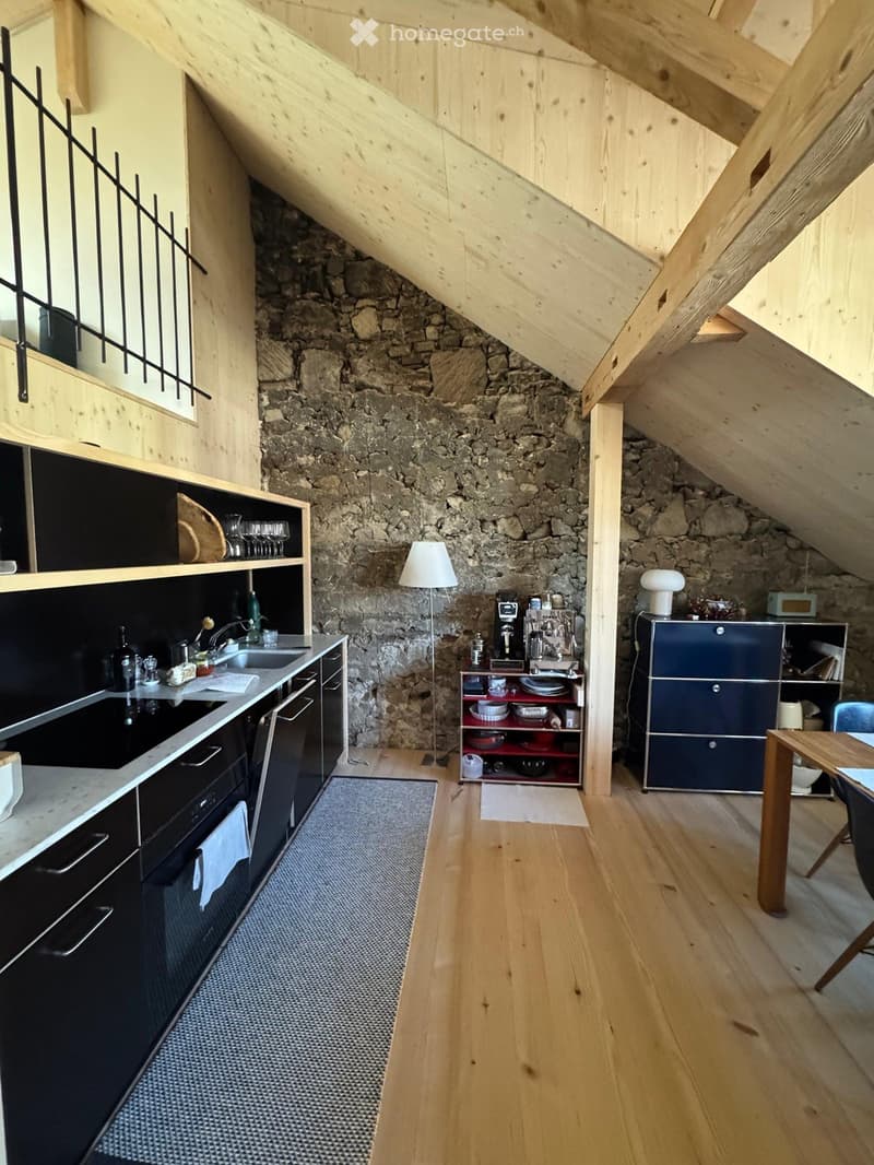 Modern kitchen with wooden floors, stone accent wall, black cabinetry, stainless steel appliances, and a small dining area.