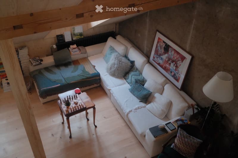 A cozy living room with a white sectional sofa, a small wooden table with a chessboard, a floor lamp, and a picture frame on the wall. The room features wooden flooring and a slanted wooden ceiling.