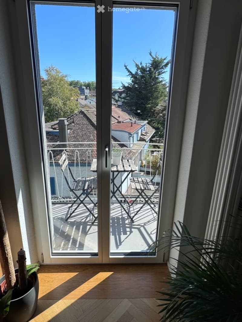 Balcony with outdoor furniture including a table and chairs, overlooking a residential area with trees and rooftops.