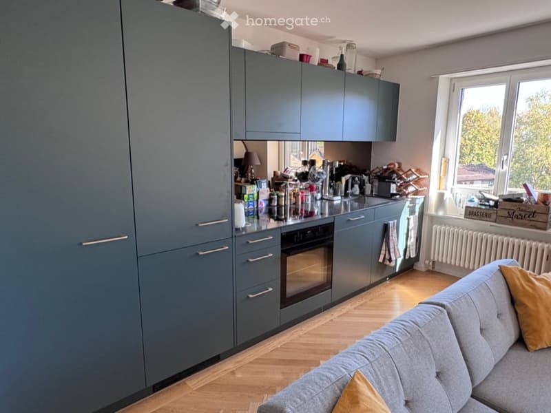 A modern, open-plan living and kitchen area with grey cabinets, a sofa with yellow pillows, and various kitchen appliances including a coffee machine and oven. The space features wooden flooring and large windows with a view of greenery.