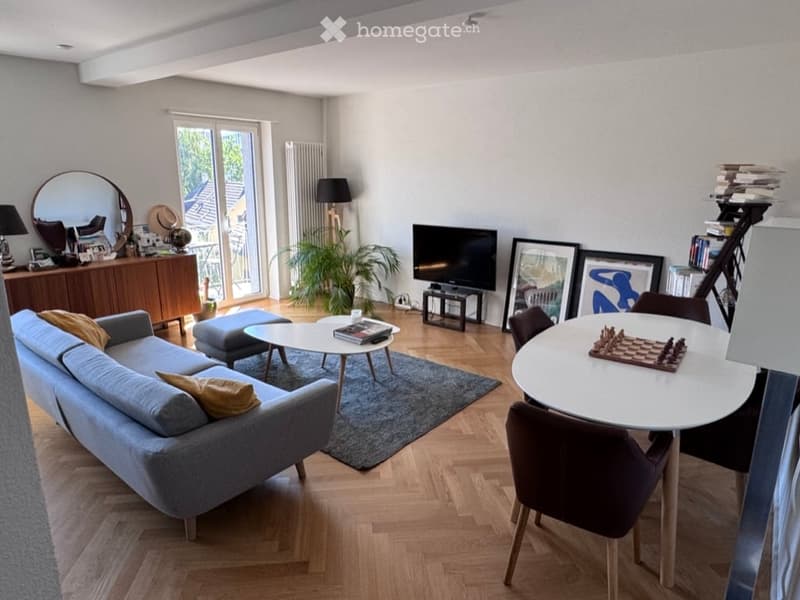 Modern living room with a grey sectional sofa, coffee table, ottoman, dining table with chairs, chessboard, mirror, lamp, plant, television, picture frames, and a bookcase.