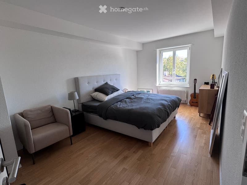Modern bedroom with wooden flooring, large window, grey upholstered bed, sofa, nightstand, lamp, guitar, and cabinet.