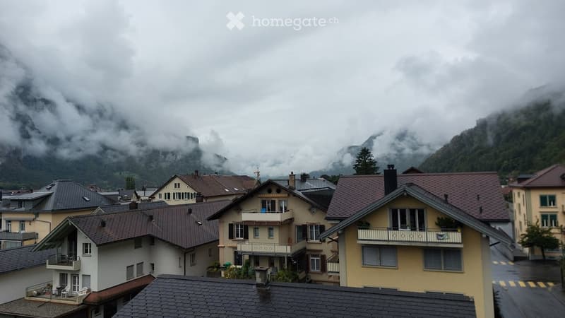 A cluster of residential buildings with sloped roofs and balconies, nestled in a mountainous valley under a cloudy sky.