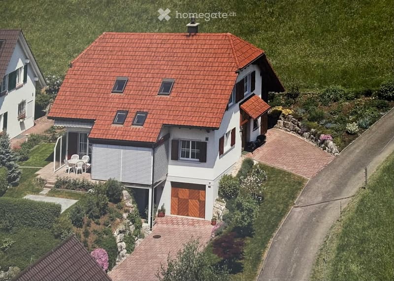 A two-story house with a red-tiled roof, white walls, and multiple windows. Features include a garage, a small patio with outdoor furniture, and a landscaped garden with pathways and greenery.