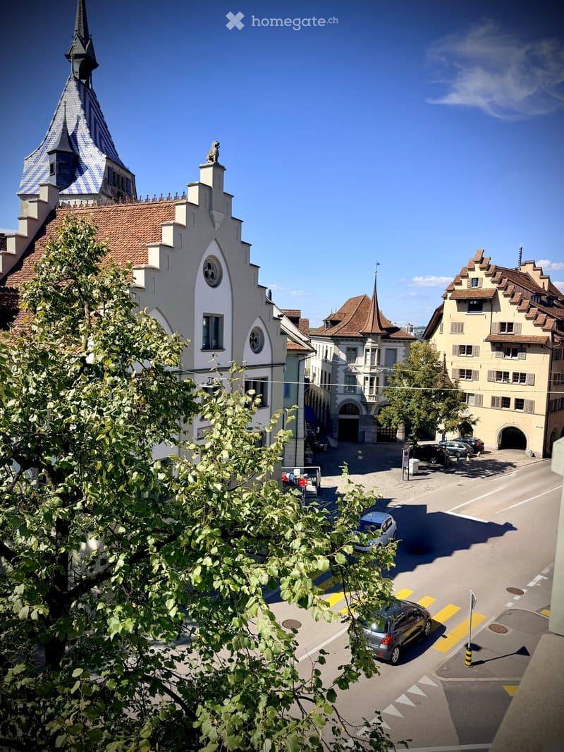 View of a historic building with a distinctive roof and spire, surrounded by other traditional European-style houses and a street with parked cars.