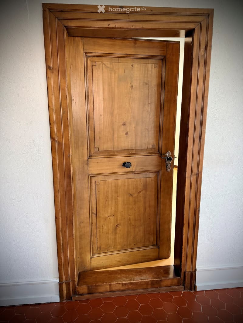Wooden door with ornate handle and lock, set in a white-walled corridor with red hexagonal tiles on the floor.