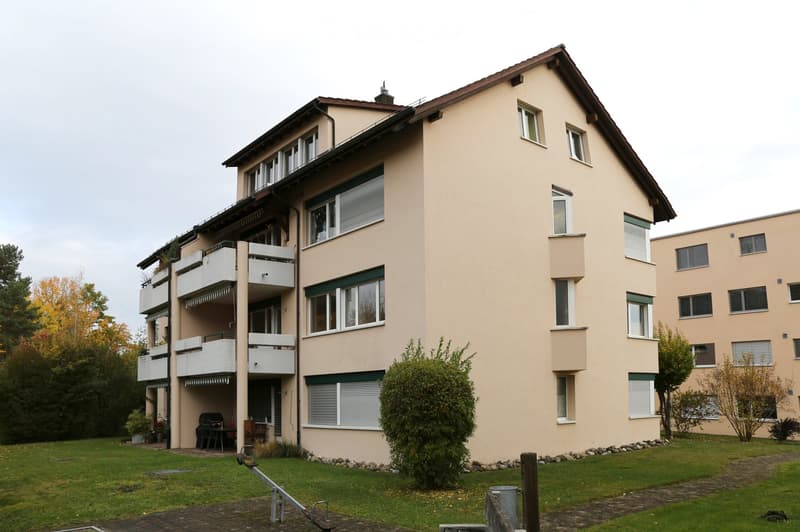 A multi-story residential building with beige walls, multiple windows, and balconies. The building is surrounded by a well-maintained lawn and trees.