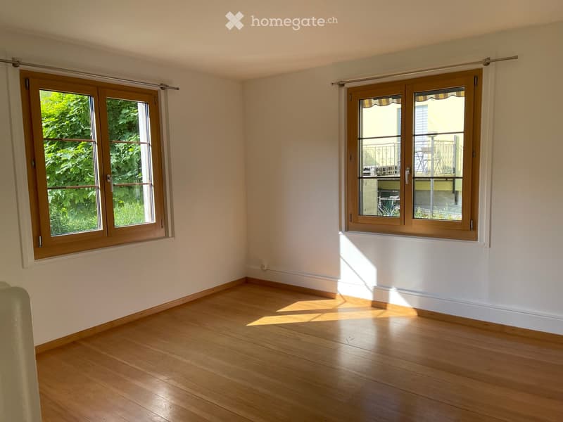 Empty room with wooden flooring, white walls, and large windows with wooden frames. Sunlight streams in, casting shadows on the floor.