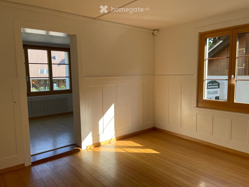 Empty room with wooden flooring, white walls, and large windows allowing natural light to enter. The room is unfurnished and has a door leading to another space.
