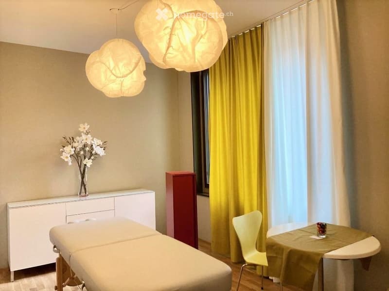 A modern, well-lit room with a massage table, a white cabinet, a small table with a green tablecloth, a yellow chair, and a vase with white flowers. The room features large windows with yellow curtains and modern pendant lights.