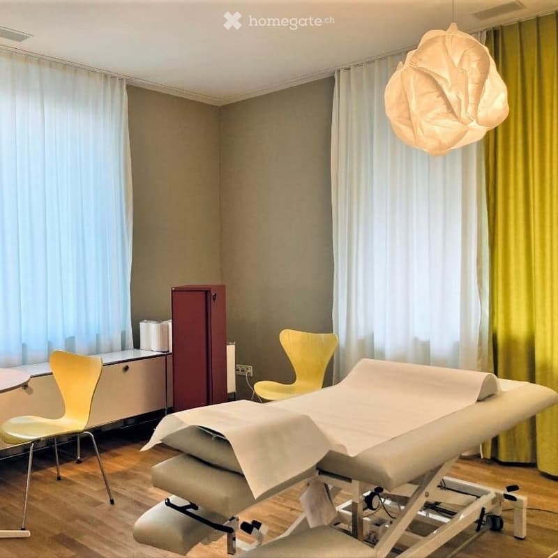 A clean, well-lit room with a massage table, two yellow chairs, a small table, and a cabinet. The room features large windows with sheer curtains and a modern pendant light.