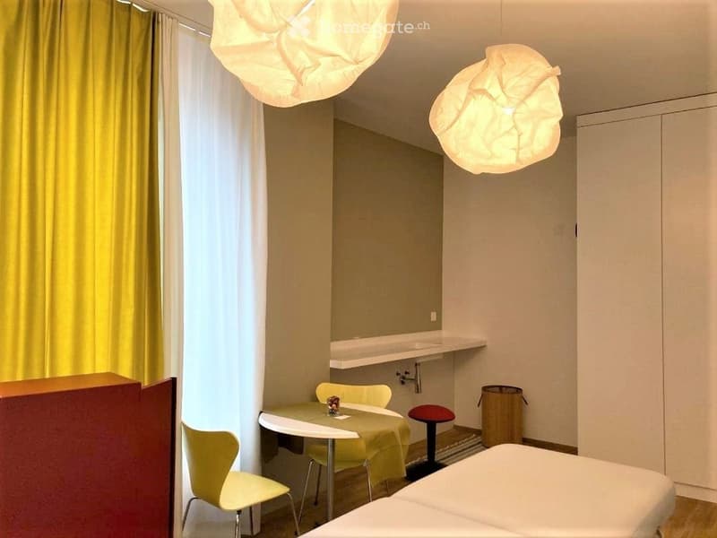 A modern, compact bedroom with a bed, small table, chairs, and a stool. The room features large windows with yellow curtains, modern pendant lights, and a small sink area with a shelf.