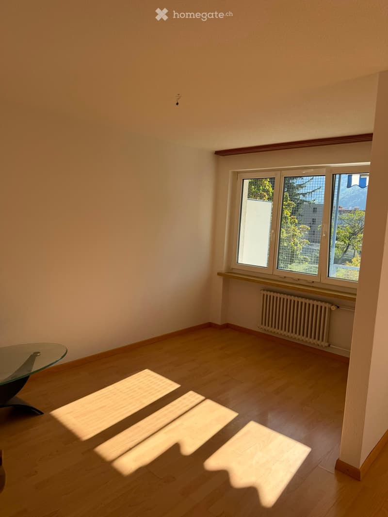 Empty room with wooden flooring, large windows, radiator, and a glass table in the corner.