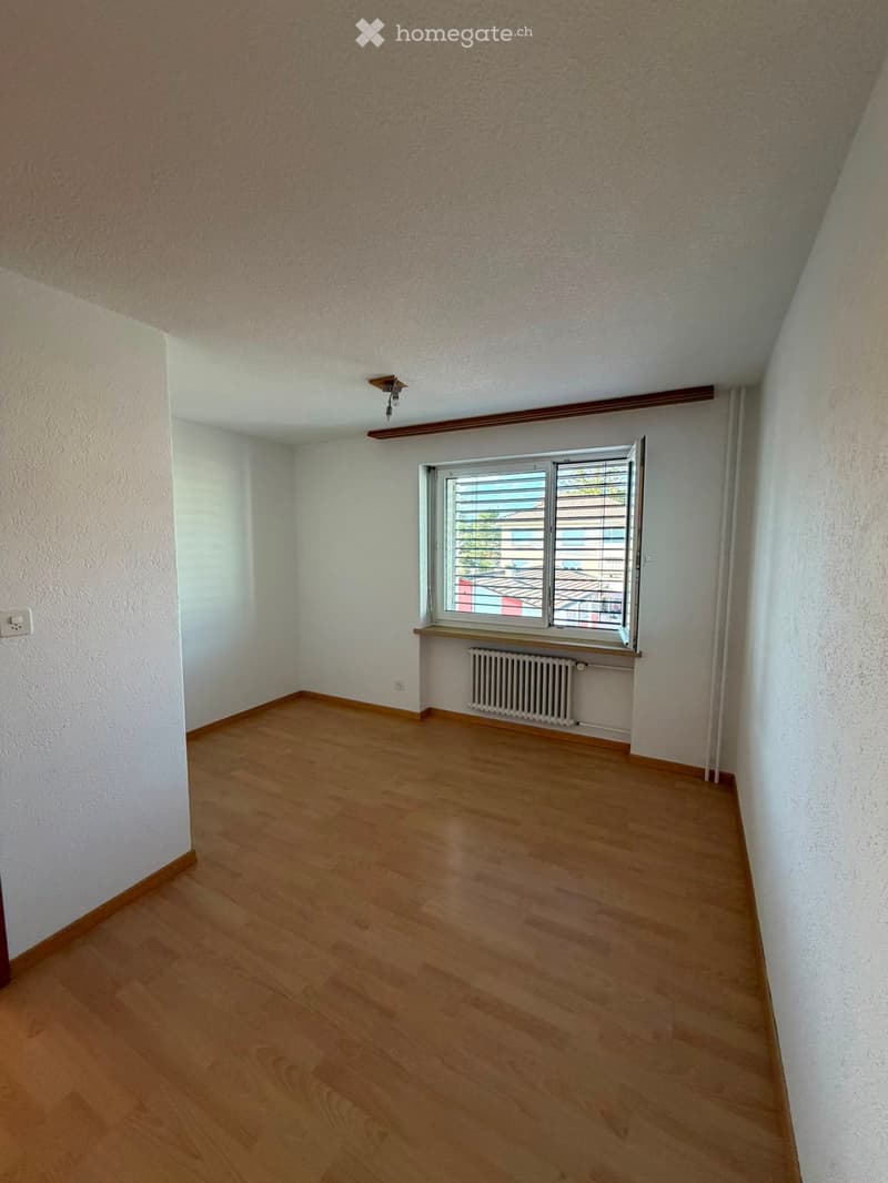Empty room with wooden flooring, white walls, and a window with blinds. A radiator is placed below the window, and the ceiling has a light fixture.