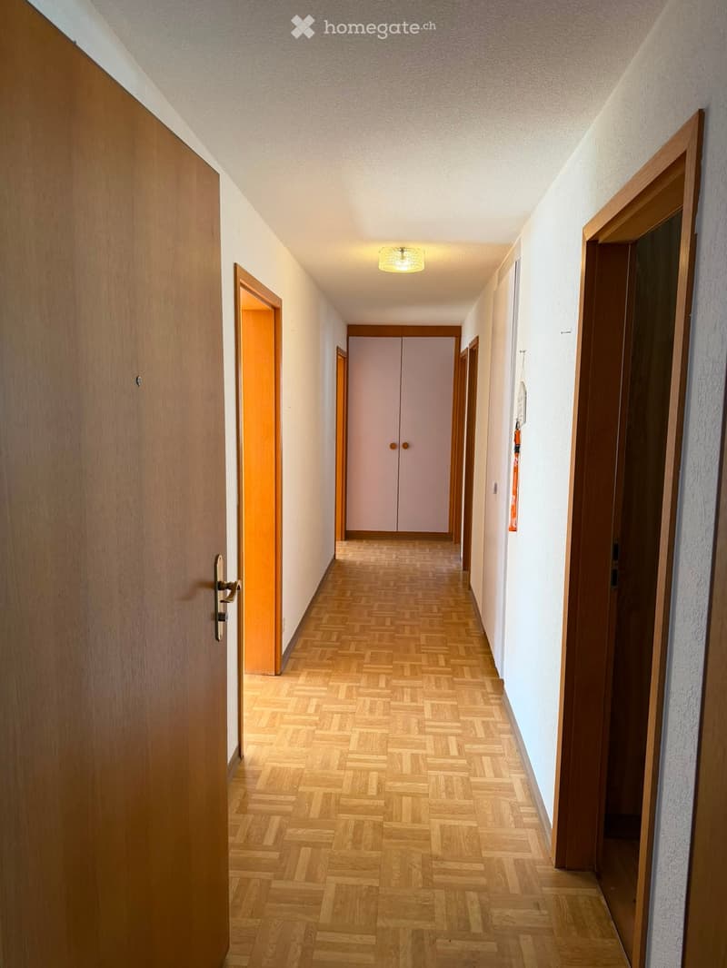 A narrow hallway with wooden flooring, white walls, and several doors, including a closet with double doors and a light fixture on the ceiling.