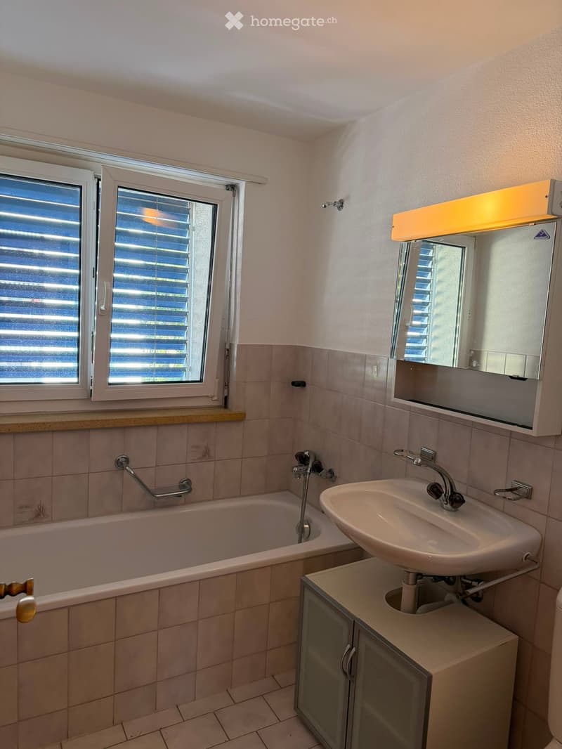 Bathroom with a bathtub, sink, mirror, and cabinet. Features tiled walls and a window with blinds.