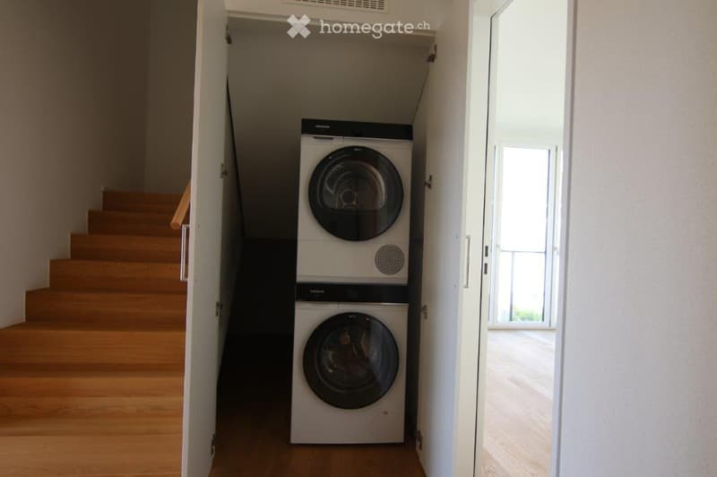 A narrow hallway with wooden flooring leads to a staircase and a private washing machine area.