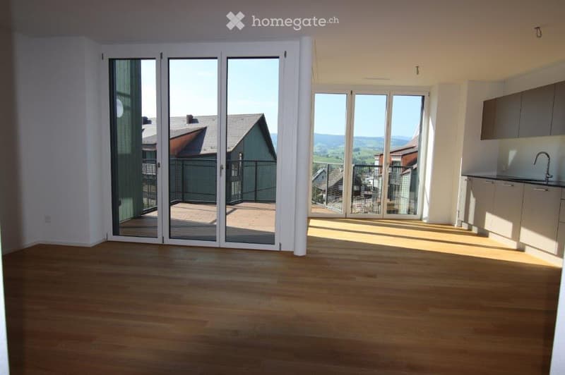 Bright, unfurnished room with wooden flooring, large sliding glass doors leading to a balcony, and a view of the surrounding landscape.