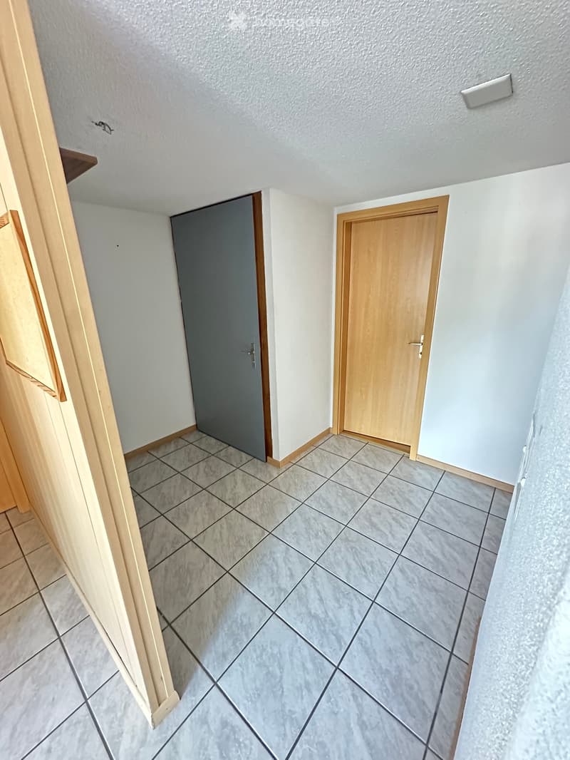 Empty hallway with tiled floor, light-colored walls, and two doors (one wooden, one grey).