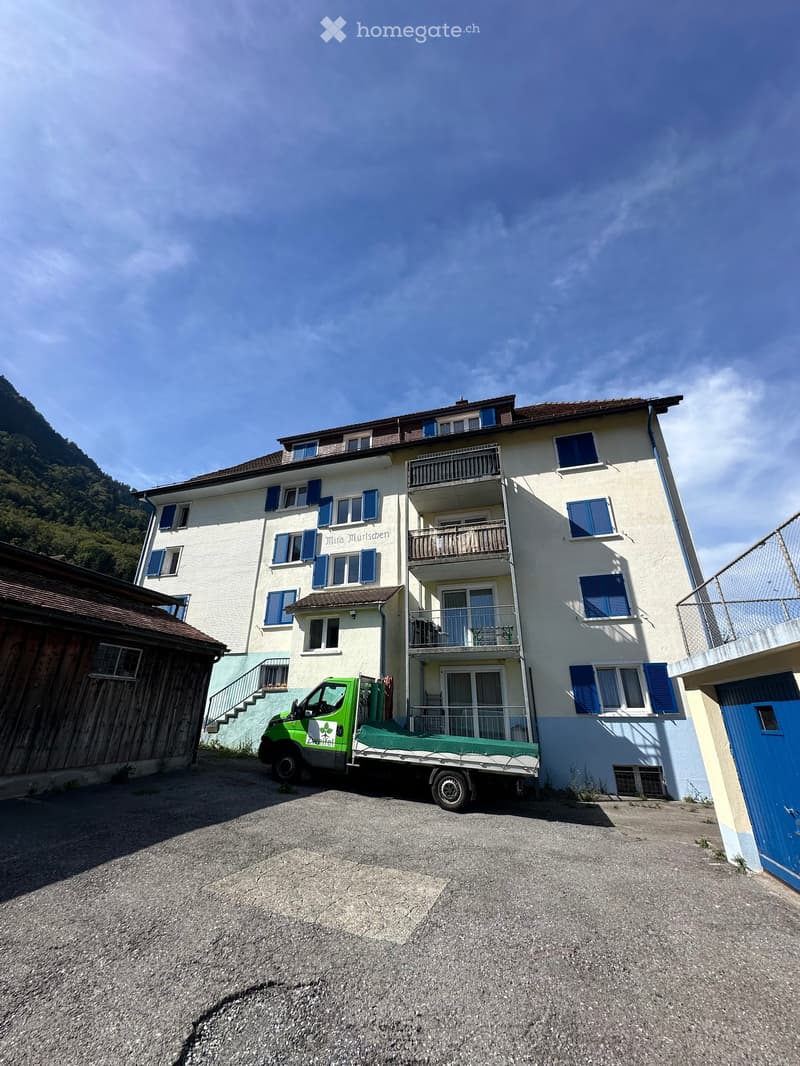A multi-story residential building with white walls, blue shutters, and balconies. A green truck is parked in front of the building.