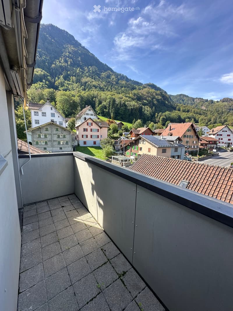 Balcony with tiled floor, overlooking a village with houses and a forested mountain backdrop.