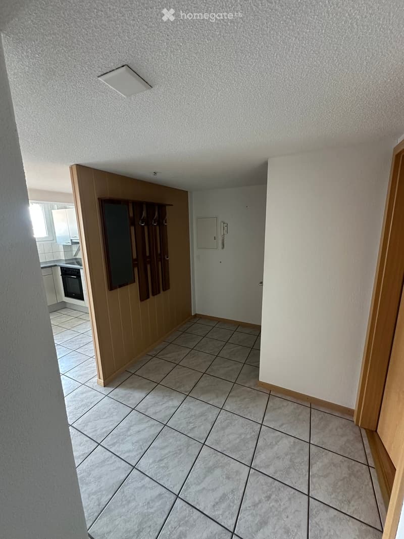 Empty hallway with tiled floor, white walls, and a wooden coat rack. A door leads to a kitchen area with visible appliances.