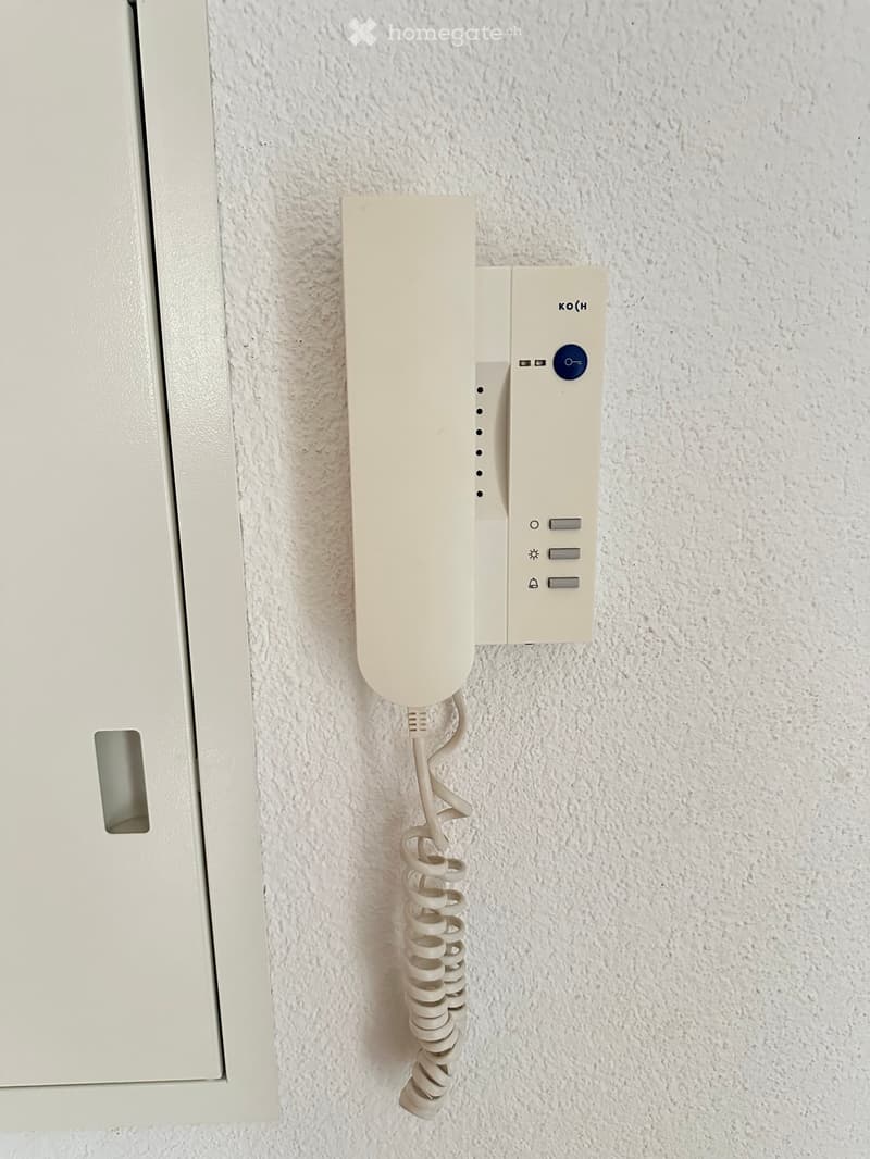 A modern intercom system mounted on a textured white wall, featuring a sleek design with a blue button and several smaller controls.