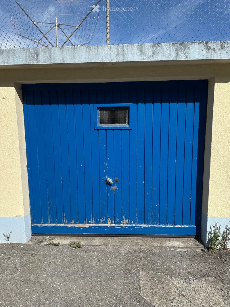 A blue garage door with a small window and a metal latch, set into a light yellow building wall with a concrete base and a chain-link fence above.