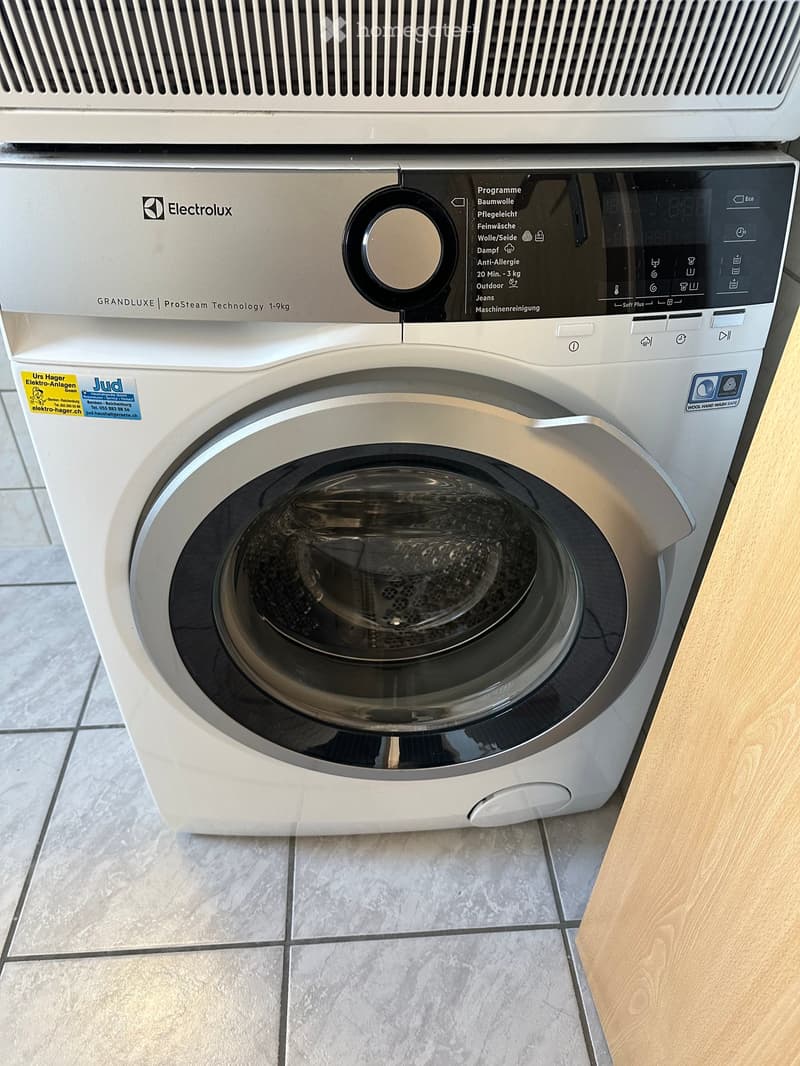 A modern washing machine with a digital control panel and a transparent door, placed on a tiled floor next to a wooden cabinet.