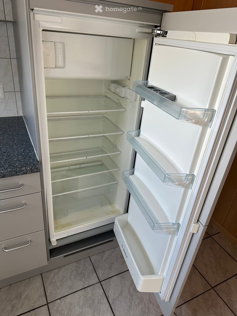 Open white refrigerator with multiple shelves and compartments, located in a modern kitchen with tiled flooring and grey cabinets.
