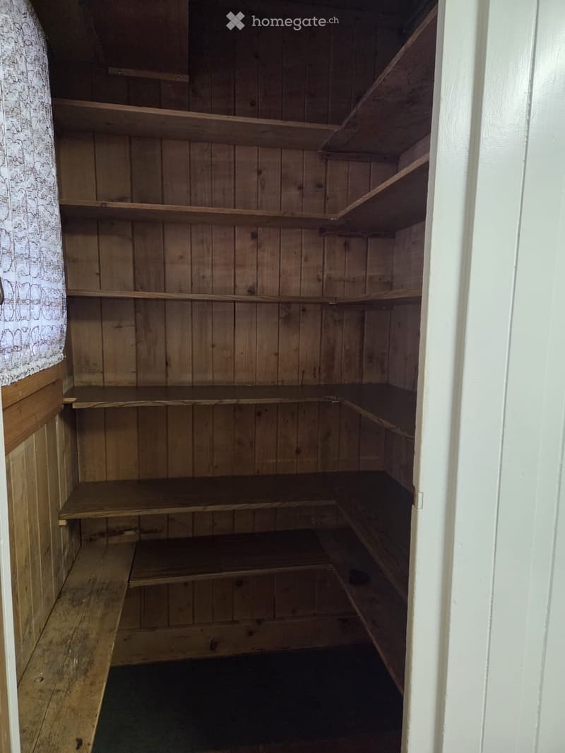 Empty wooden closet with multiple shelves and a small bench at the bottom, featuring wooden walls and a white door frame.