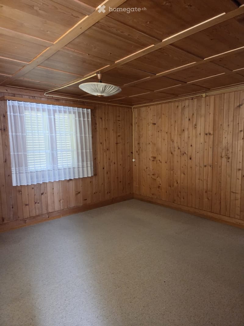 Empty room with wooden walls and ceiling, carpeted floor, and a single hanging light fixture. White curtains cover a window on one wall.