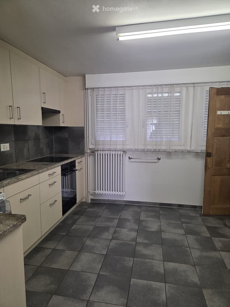 Empty kitchen with light-colored cabinets, granite countertop, stainless steel sink, and tiled floor. Window with blinds and radiator visible.