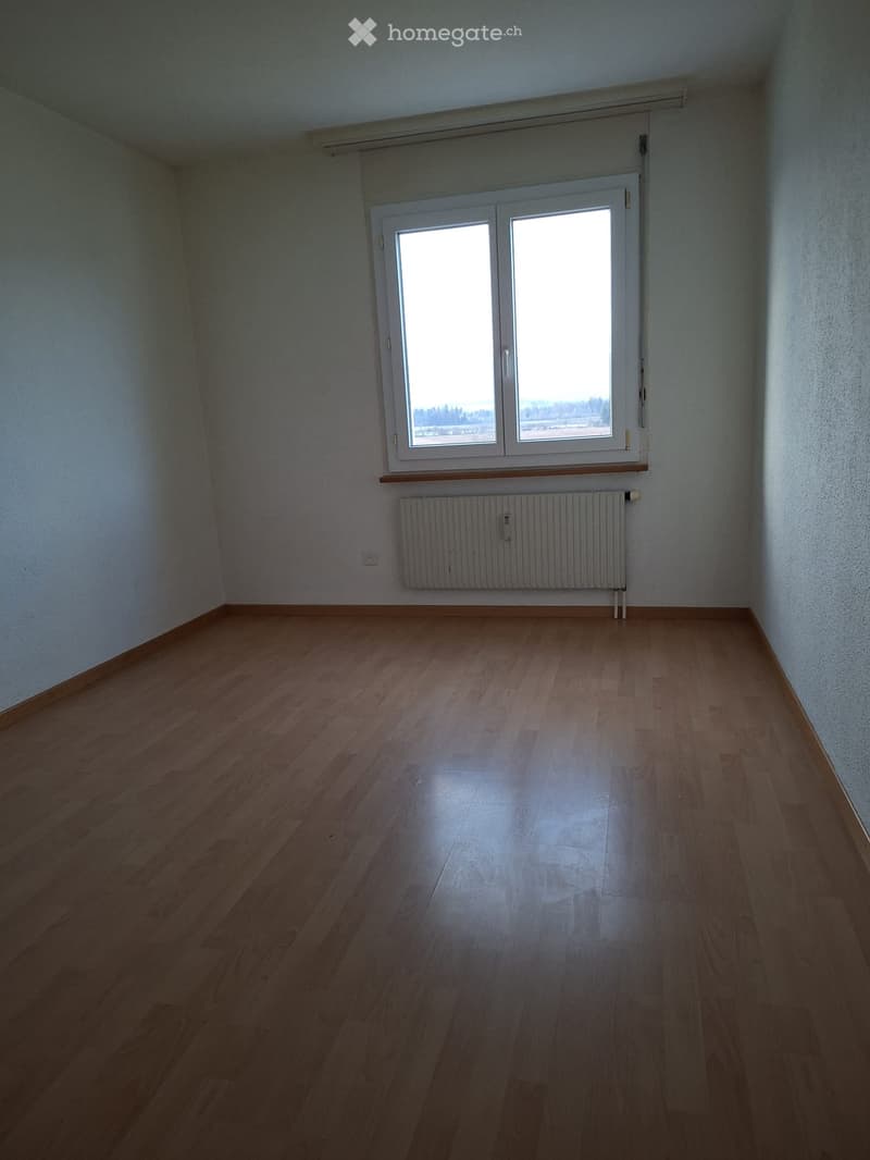 Empty room with wooden floors, white walls, large window, and radiator