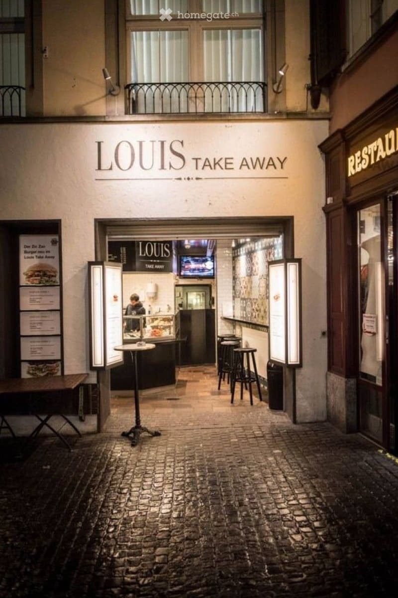 Facade of a restaurant named "Louis Take Away" with a small outdoor seating area featuring tables and chairs, illuminated signage, and a balcony above.