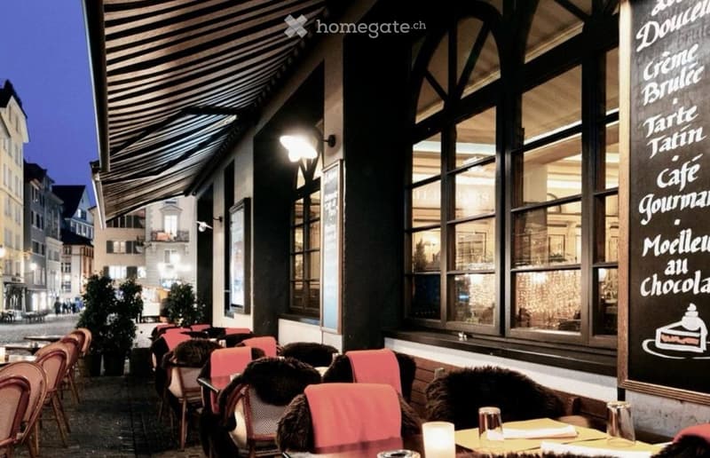 Outdoor seating area of a restaurant with red chairs, tables, and potted plants, illuminated by streetlights.