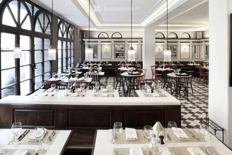 Elegant restaurant interior with large arched windows, checkered flooring, multiple dining tables set with wine glasses, cutlery, and napkins, and a central bar counter.