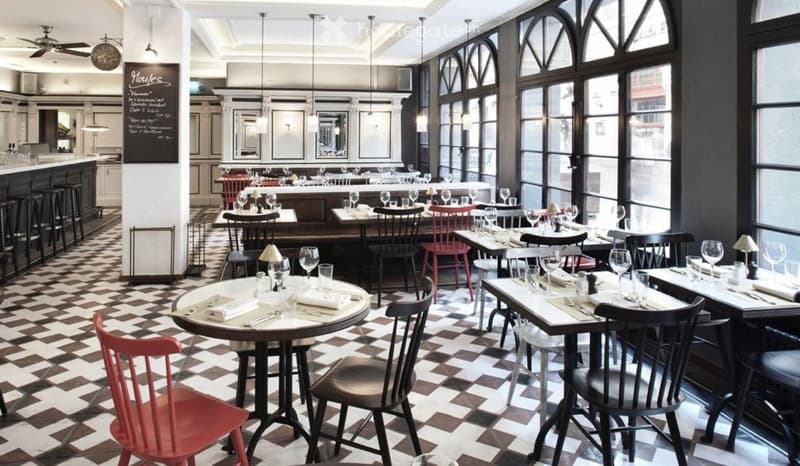 Restaurant interior with checkered flooring, large windows, round tables with wine glasses, and red and black chairs.