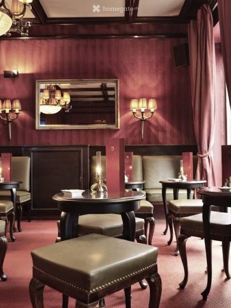 Elegant lounge area with dark wood furniture, plush leather stools, a sofa, small tables, wall-mounted lamps, and a decorative mirror.