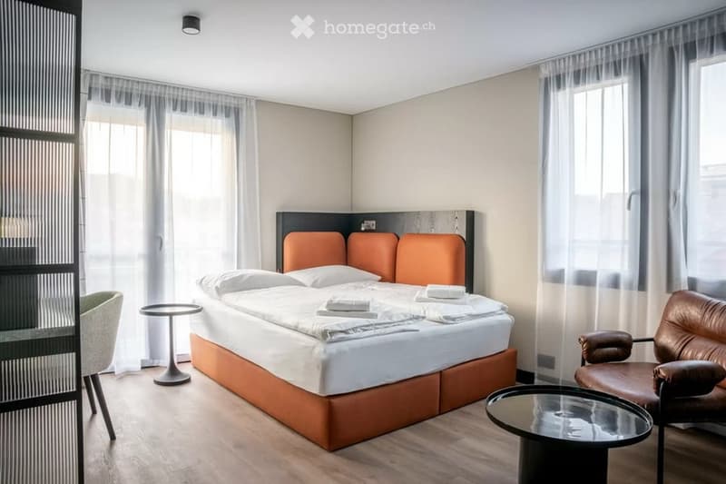 Modern bedroom with a large bed, orange headboard, two chairs, and a small table. The room features large windows with sheer curtains and a sleek, contemporary design.