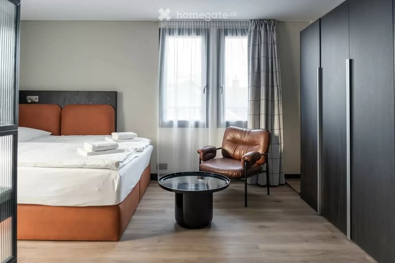 Modern bedroom with a large bed, leather chair, round glass table, and large windows with curtains.
