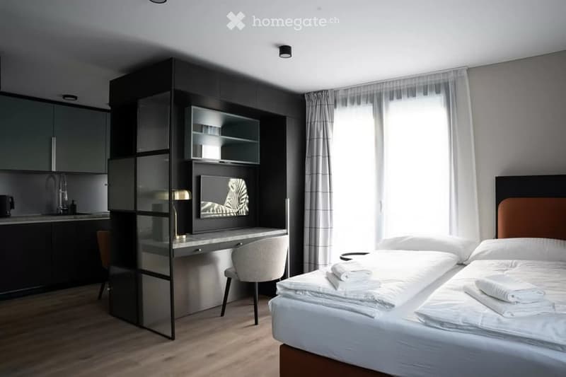 Modern, well-lit bedroom with a large bed, a desk with a chair, a mirror, and a small refrigerator. The room features wooden flooring and large windows with sheer curtains.