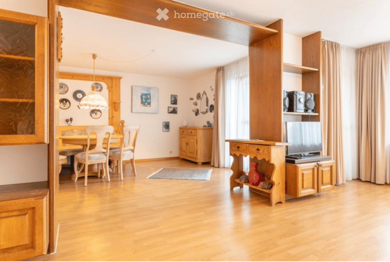 Bright, open-plan living and dining area with wooden flooring, large windows, and natural light. Features a dining table with chairs, a wooden cabinet, a television on a stand, a floor lamp, and decorative picture frames on the wall.